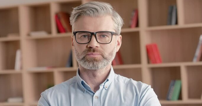 Portrait of Caucasian man with grey hair and beard. Male wearing glasses and blue shirt. Glancing at camera with serious look. Confident person. Standing in library or office.