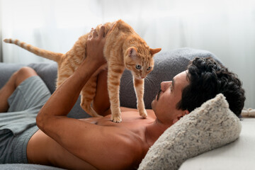 Brown tabby cat  on the chest of a young man lying on a sofa. The man strokes the cat's head . close up - Powered by Adobe