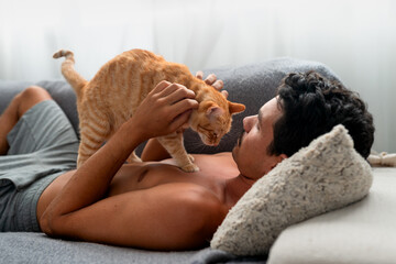 Brown tabby cat  on the chest of a young man lying on a sofa. The man strokes the cat's head ....