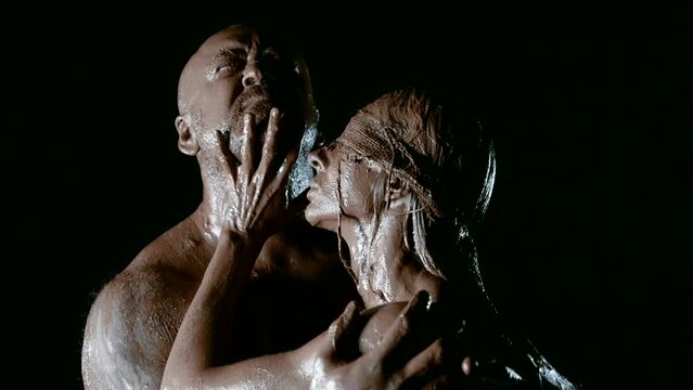 On a black background in the darkness of the night, a strong-built man and his clay-covered wife share an unbreakable bond and deep love.