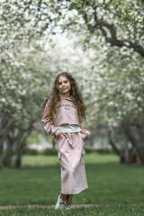 A beautiful toddler girl in a pink dress stands by a blooming white apple tree in the park and enjoys the beauty of a blooming garden, spring.