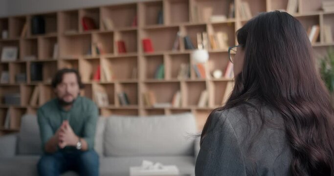 In blurred vision Caucasian male telling something actively. Psychologist in focus sitting in front of him. Disagreeing by nodding her head. Man with active hand movements describing moment.