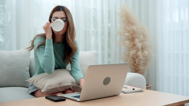 A young woman happily sits on a sofa using a laptop to do online work at home.