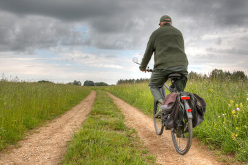An active senior rides his bicycle on a dirt path between two green meadows. The sky is cloudy and...