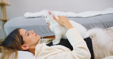 Attractive woman is laying on a bed petting cute little fluffy white cat in living room, owner...