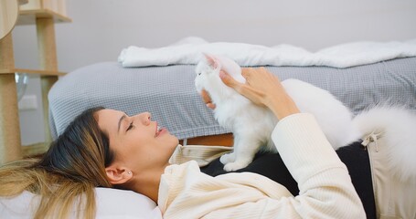 Attractive woman is laying on a bed petting cute little fluffy white cat in living room, owner...