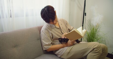 Asian man sitting on a couch stroking lovely fluffy cat with hand on his lap while reading a book...