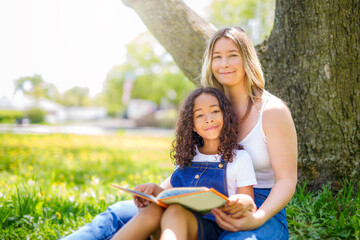 Black American child is reading book with her mother while having a summer picnic in the public...