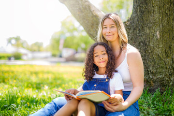 Black American child is reading book with her mother while having a summer picnic in the public park.