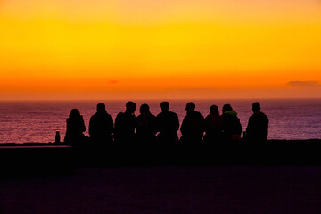 Silhouettes of young people sitting in front of a sea with an orange sunset sky with the sun setting 