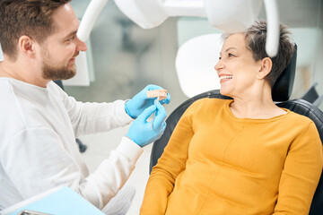 Cheerful aesthetic dentist showing to client how he treat her tooth