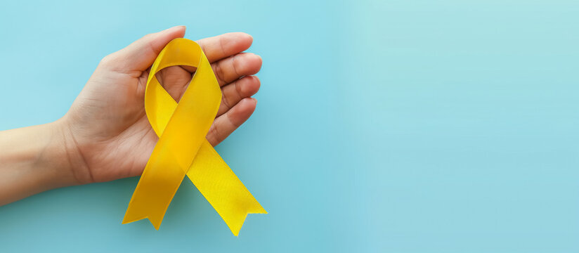 hand holding a yellow ribbon for cancer awareness on a light blue background with copy space for text