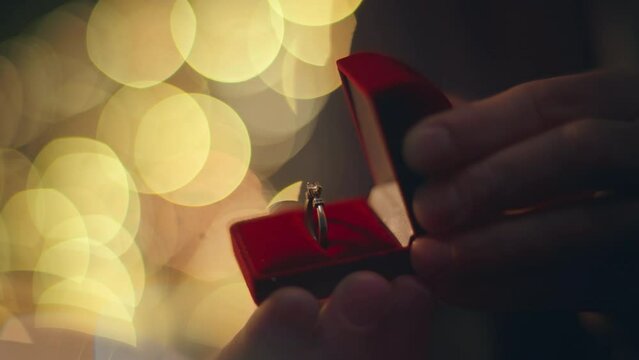 Groom hands opening ring red box at evening lights blurred background closeup