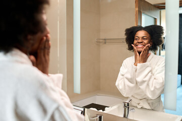 Portrait of smiling young Black woman looking in mirror enjoying skincare in morning, copy space