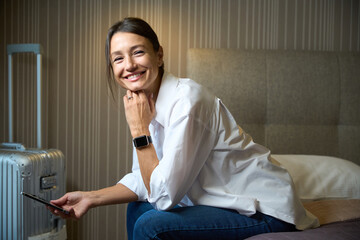 Side view of young smiling woman with smartphone looking at camera on bed