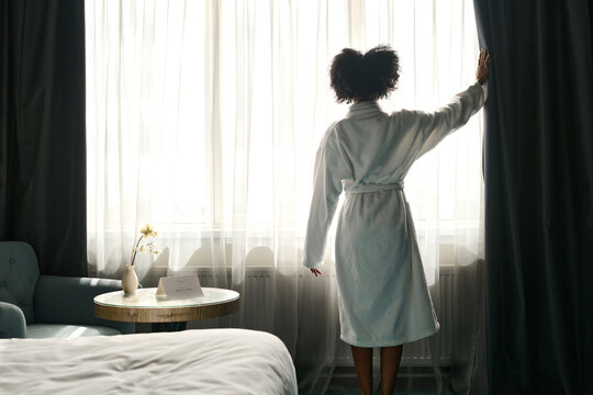 Back view of African American woman wearing bathrobe enjoying morning in hotel room and opening window curtains, copy space - Powered by Adobe