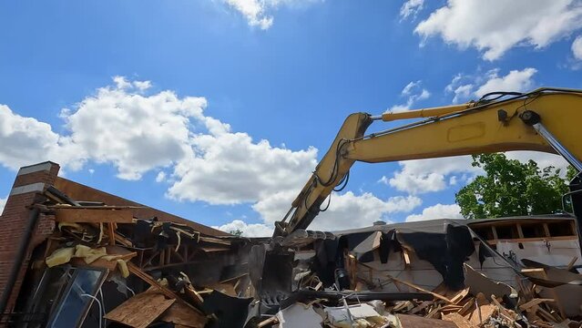 Hydraulic excavator demolishes a building. Industrial equipment with large claw destroys the side of a brick building.  Sunny skies and clouds above the scene. Slow motion views.