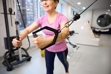 Fototapeta premium Obscure face of mature woman doing exercise for chest muscles in blurred gym