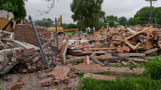 Small loader tractor equipment moves in reverse through a demolition site. Piles of rubble in the foreground of view following destruction of a building. Overcast cloudy skies above.
