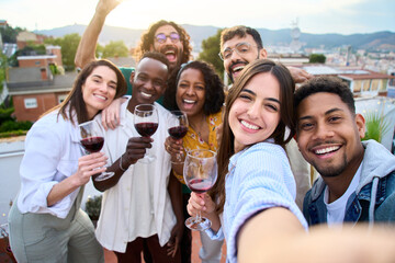 Group of diverse friends taking selfie and having fun at rooftop home party holding glass of red...