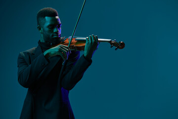 Fototapeta premium Elegant African American man in suit playing violin against blue background in studio setting