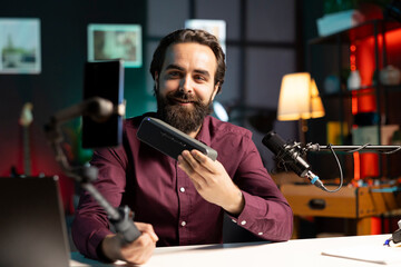 Cheerful online star doing technology reviews, excited to look at mini soundbar specifications. Influencer in studio delighted by high end music playing device, filming with smartphone on selfie stick