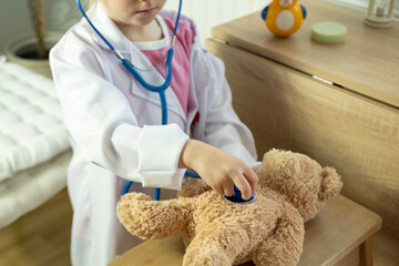 little child, blonde girl 4 years old plays with toy, little doctor carefully checking teddy bear's heartbeat with stethoscope, vocational guidance, plush friend