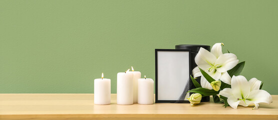 Blank funeral frame, burning candles and beautiful lily flowers on wooden table near color wall