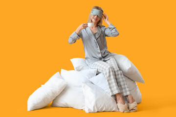 Pretty young woman in pajamas and with coffee sitting on pillows against yellow background