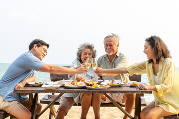 Happy Asian adult couple and aging parents enjoy outdoor lifestyle celebration dinner party drinking champagne together at tropical beach restaurant during travel ocean on summer holiday vacation. 