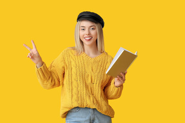 Beautiful young woman with book showing victory gesture on yellow background