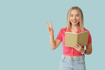 Beautiful young happy woman with book showing victory gesture on blue background