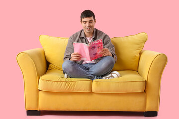 Handsome young man reading magazine on yellow sofa against pink background