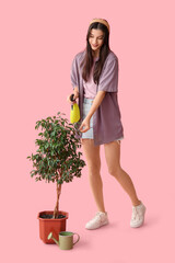 Beautiful young happy female gardener spraying water onto tree on pink background