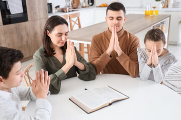 Family praying with Holy Bible on table in kitchen