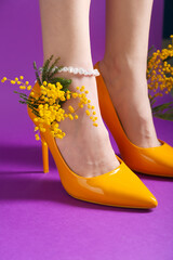 Legs of beautiful young woman in orange high heels with mimosa on purple background