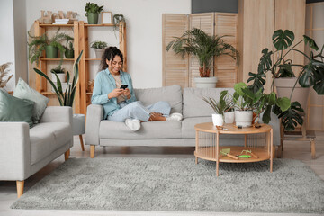 Beautiful young African-American woman using mobile phone among plants in living room