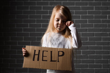 Homeless little girl holding piece of cardboard with word HELP near grey brick wall