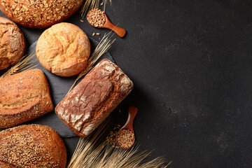 Loaves of fresh bread with buns, wheat spikelets and grains on black background