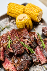 A dish of Black Angus steaks, roasted corn, rosemary and salt on a plate. Yellow baked corn cobs. Sprigs of rosemary.