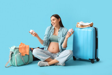 Young pregnant woman with baby booties, maternity hospital bag and suitcase sitting on blue...