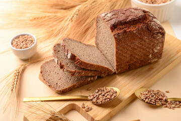 Wooden board with sliced loaf of bread, wheat spikelets and grains on beige background