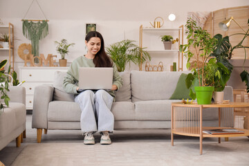 Beautiful young woman with modern laptop and houseplants sitting on sofa in living room