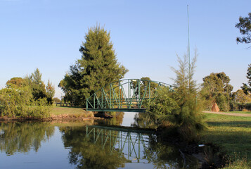 Fototapeta premium Green metal bridge over a lake of water surrounded by trees and plants at McCaughey Bicentennial Park in Yanco, New South Wales, Australia