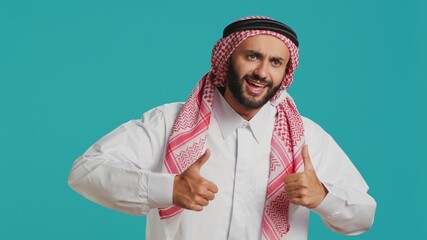 In front of camera, middle eastern guy gives thumbs up indicating consent and fulfillment while dressed in customary muslim apparel. Young person wearing traditional arab clothing, like symbol.