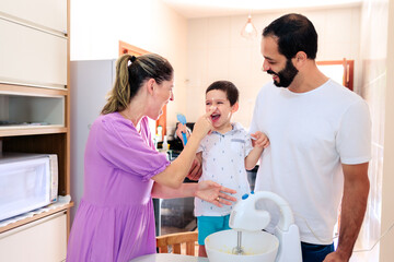 mom, dad, and young son making a cake together. Mom playfully puts cake batter on her son's nose, making everyone laugh. The image captures love and joyful family moments.