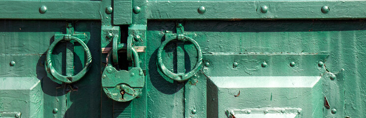 The metal gate is padlocked. Rusty padlock on the gate, close-up. Rough iron doors painted green.