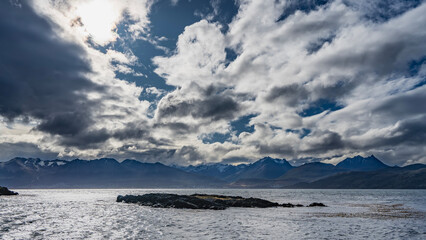 A small rocky islet in the ocean. Ripples, algae on the surface of the water. A picturesque snow-capped mountain range of the Andes against a blue sky, clouds. Argentina. Patagonia.The Beagle Channel