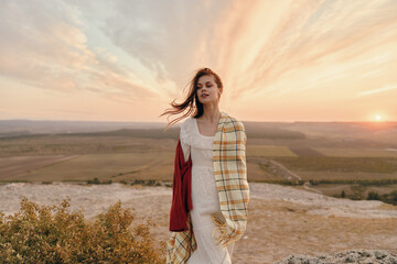 Woman in white dress and plaid scarf standing on top of hill at sunset with travel and beauty theme