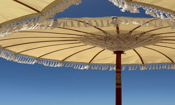 Relaxing video footage of a bohemian beach parasol gently swaying in the sea breeze against a backdrop of blue sky in Bali, Indonesia.